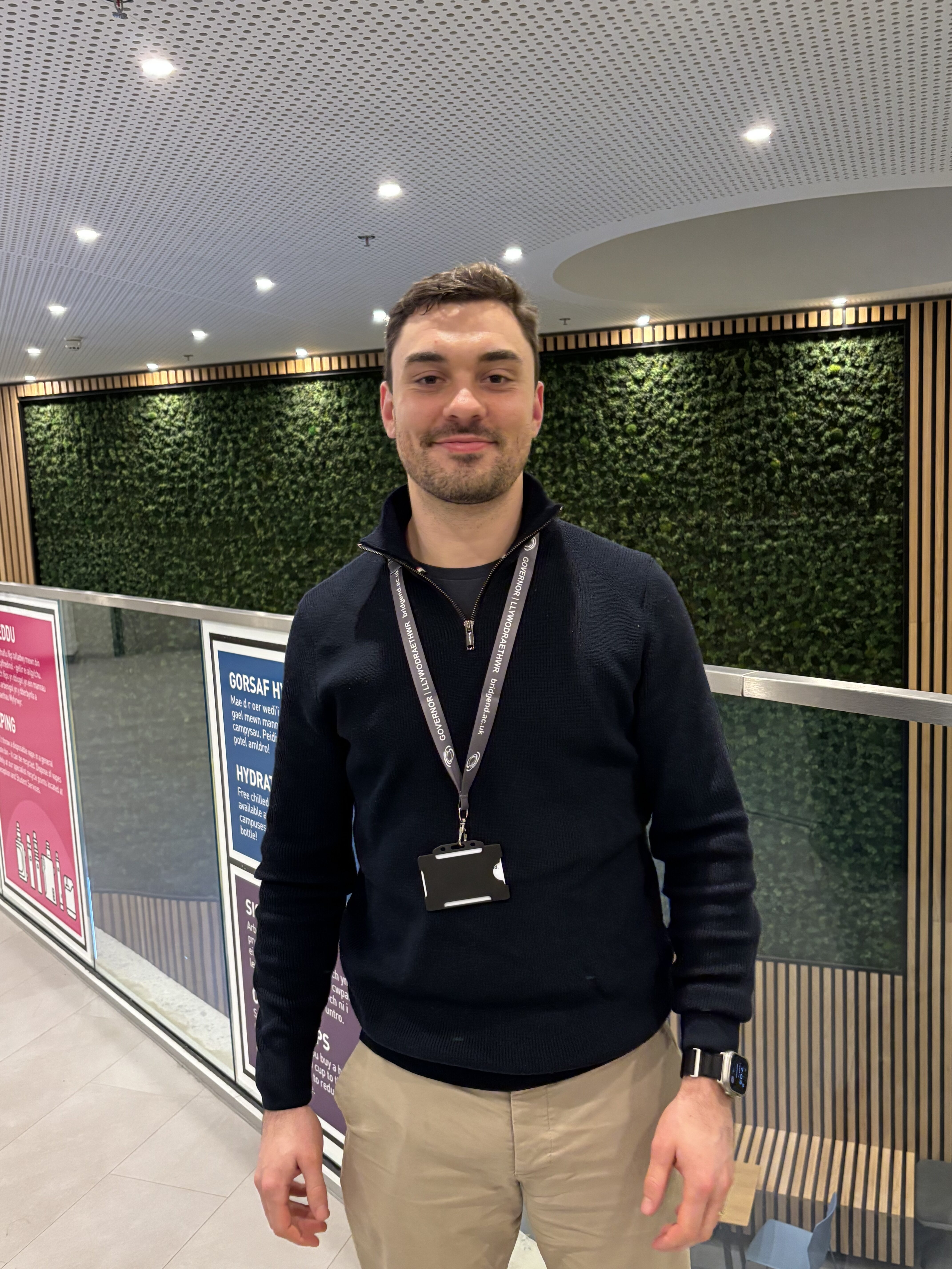 A smiling man in a dark jumper and lanyard standing in a modern building with a green foliage wall background.
