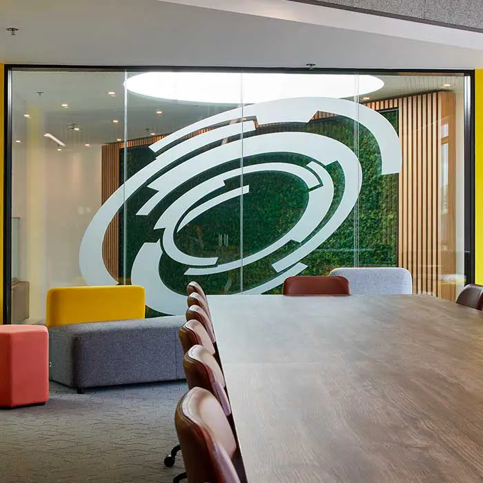 A wooden meeting table surrounded by chairs, facing glass partition walls with a circular graphic and yellow accents.