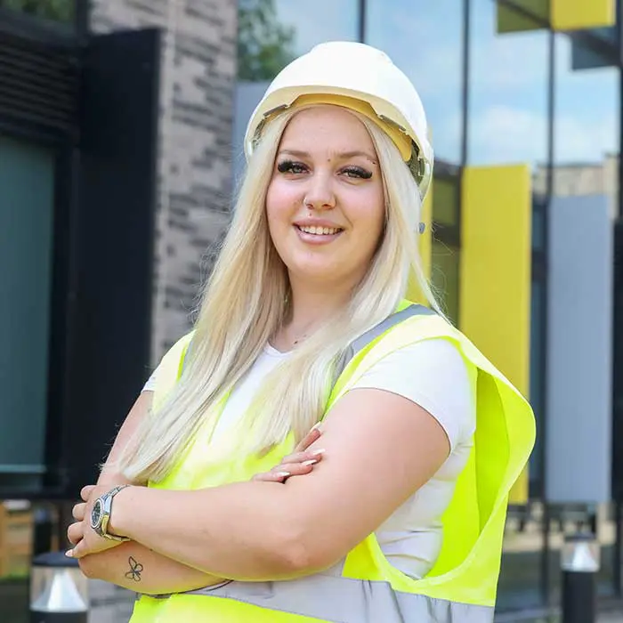 A smiling woman wearing a white hard hat and high-vis vest standing with folded arms outside a modern building.