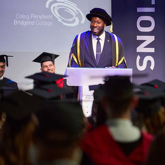 A smiling speaker in academic regalia giving a speech at a lectern during a Bridgend College graduation ceremony.